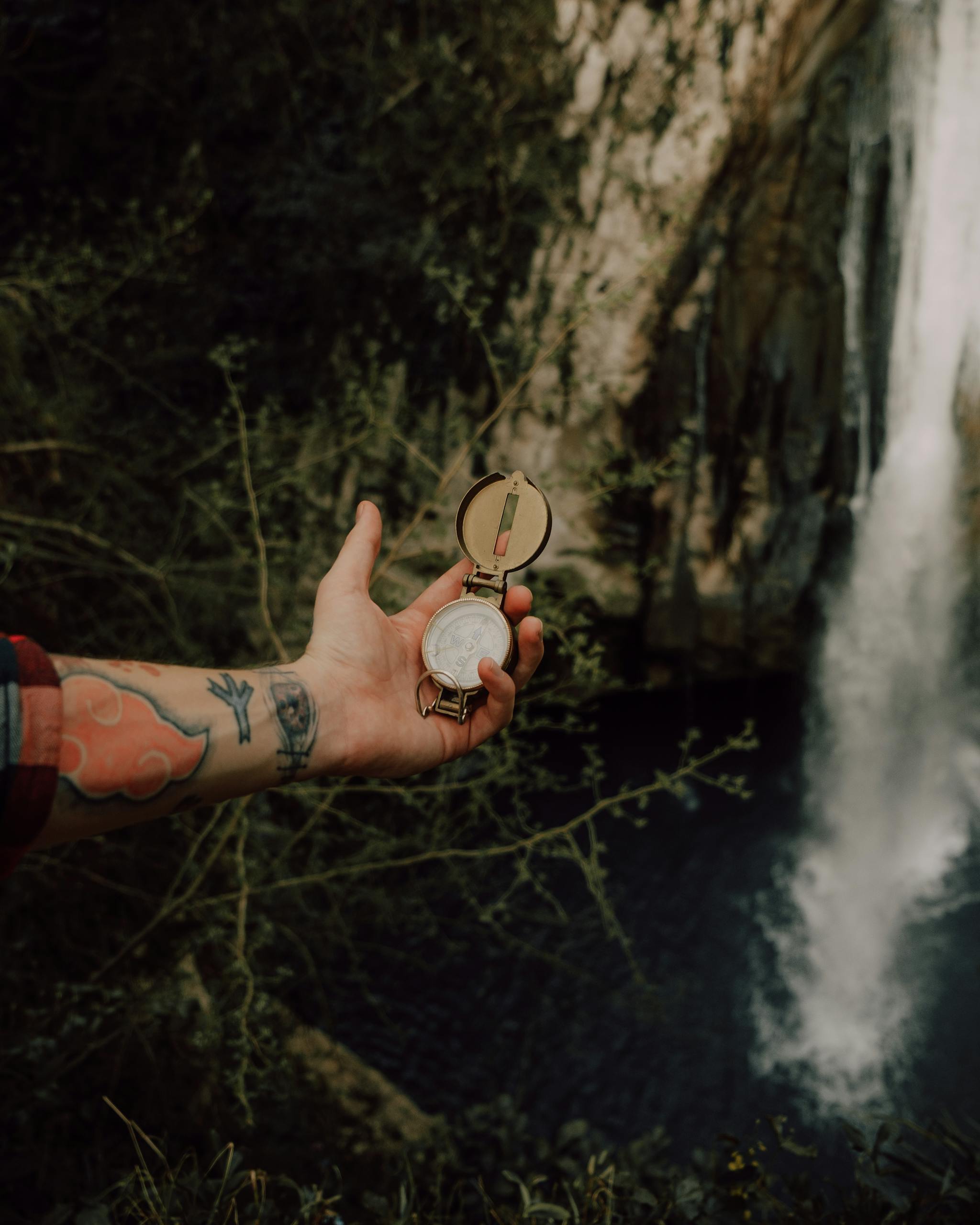 A tattooed arm holding a compass near a waterfall, surrounded by lush forest scenery.